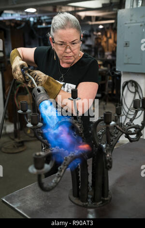 Caucasian female blacksmith using a torch to heat a metal candelabra in her studio. Stock Photo
