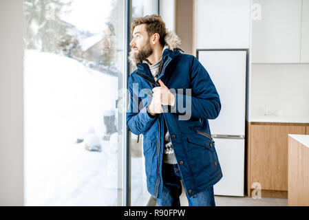 Man dressed in winter clothes feeling cold standing near the window at home with no heating Stock Photo