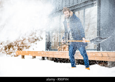 Handsome man in winter clothes cleaning snow with a shovel near the modern house in the mountains Stock Photo