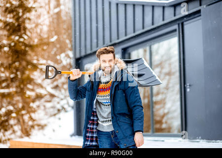 Portrait of a man in winter clothes with a snow shovel near the modern house in the mountains Stock Photo