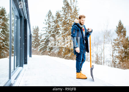 Portrait of a man in winter clothes standing with snow shovel on the terrace of the building in the mountains
