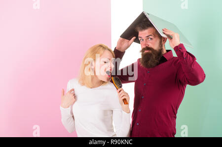 Man fed up listening her creepy voice. Crazy about her voice. Lady imagine she superstar talented singer. Lady awful voice sing using hair brush as microphone while man annoyed hiding under laptop. Stock Photo