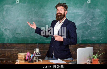 Teacher bearded man tell interesting story. Teacher interesting interlocutor best friend. Talking to students or pupils. Teacher charismatic hipster stand near table classroom chalkboard background. Stock Photo