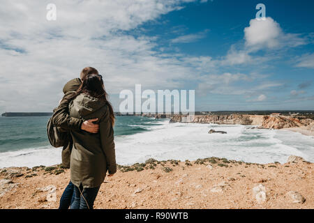 Rear view of a young couple of tourists or travelers enjoying in solitude a beautiful view of the landscape and the Atlantic Ocean in Portugal. Joint travel and pastime. The guy hugs the girl. Stock Photo