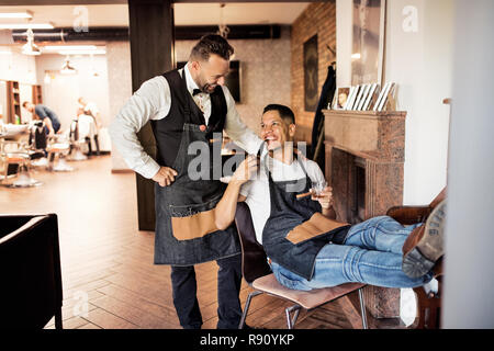 Two cheerful haidresser and hairstylist in barber shop, smoking a pipe and talking. Stock Photo