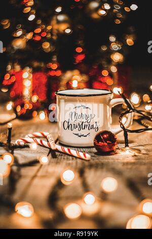 Close up of a white mug with christmas lights and christmas decorations on a wooden table with the message Merry Christmas Stock Photo