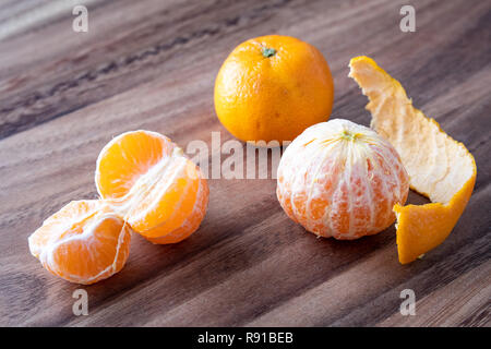 Three satsuma oranges on a rustic wood table, two oranges peeled, one sectioned, and peels on the table Stock Photo