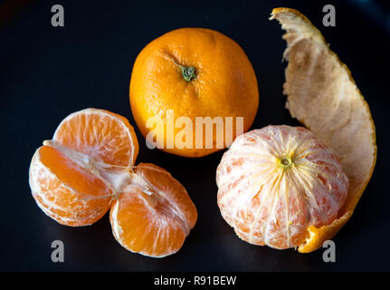 Three satsuma oranges on a black plate, two oranges peeled, one sectioned, and peels on the plate Stock Photo