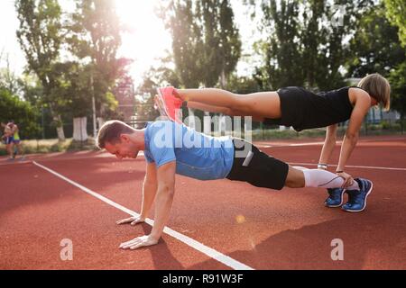 Man in blue t-shirt and black shorts standing in plank. Young woman in black sportswear standing on the man. Morning force training together. Stock Photo
