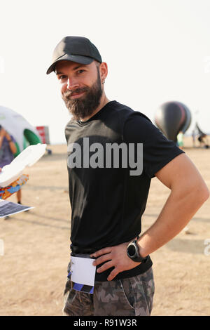 Portrait of a sports guy with the beard in black t-shirt and grey shorts, cap on the head, accessories on the arm. Standing outdoor on the field Stock Photo