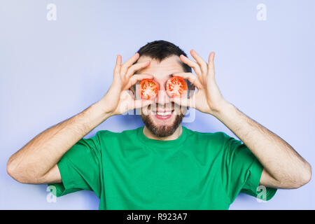 Young bearded and smiling man holding slices of red tomatoes in front of his eyes. Light purple background. Stock Photo