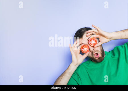 Young bearded and showing surprise man holding slices of red tomatoes in front of his eyes. Light purple background, copy space. Stock Photo