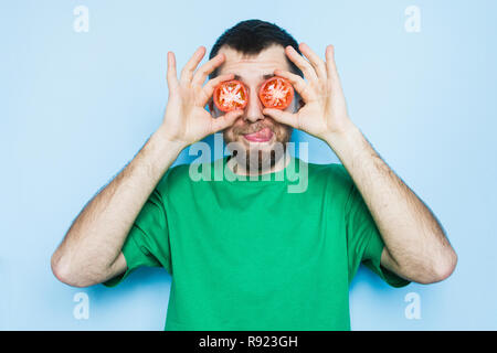 Young bearded man holding slices of red tomatoes in front of his eyes, showing his tongue. Light purple background, copy space. Stock Photo