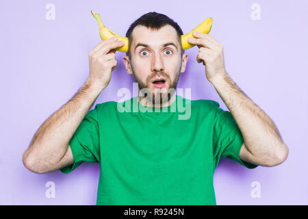 Young bearded man holding cut in half yellow banana on the level of temples, surprised and shocked face expression. Light purple background, copy spac Stock Photo