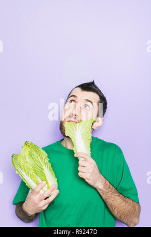 Young man biting on a leaf of Beijing napa cabbage, doubt face expression, looking up. Light purple background, copy space, vertical orientation. Stock Photo
