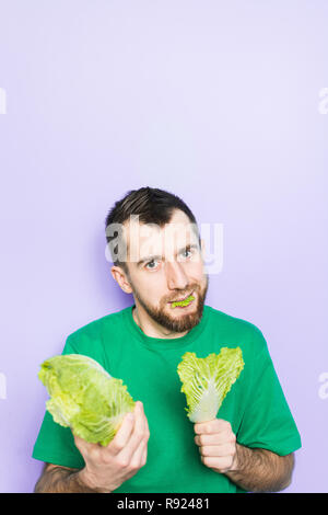 Young man biting on a leaf of Beijing napa cabbage, offering the product to the viewer. Light purple background, copy space, vertical orientation. Stock Photo