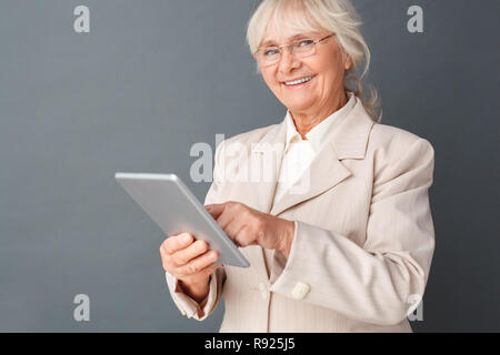 Senior woman in fromal suit and glasses studio standing isolated on gray using application on digital tablet cheerful close-up Stock Photo