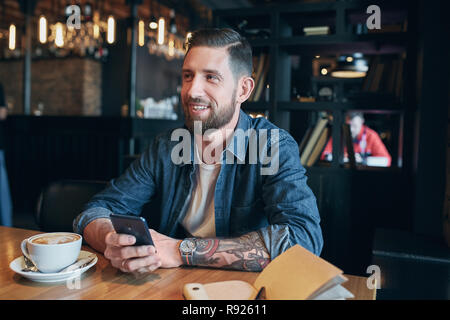Young bearded businessman,dressed in a denim shirt, sitting at table in cafe and use smartphone. Man using gadget.