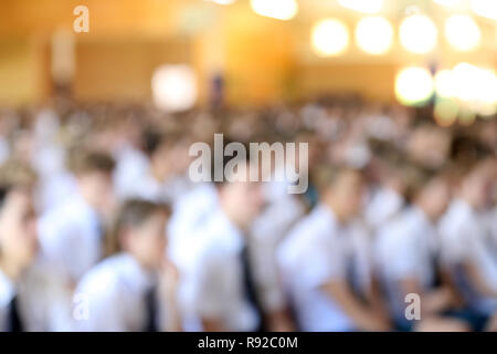 Deliberately out of focus blurred image of large hall full of high school students in white shirt and ties uniforms listening at assembly. Stock Photo