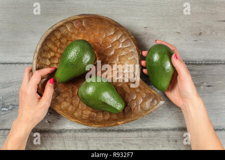 Tabletop view - woman hands holding wooden carved bowl with three green avocado pears. Stock Photo