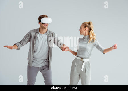 couple in grey clothing holding hands while man experiencing virtual reality isolated on grey Stock Photo