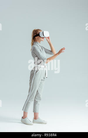 girl in grey clothing and vr headset gesturing while having virtual reality experience on grey background Stock Photo