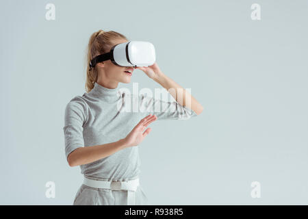 girl in grey clothing and vr headset touching air while having virtual reality experience isolated on grey Stock Photo