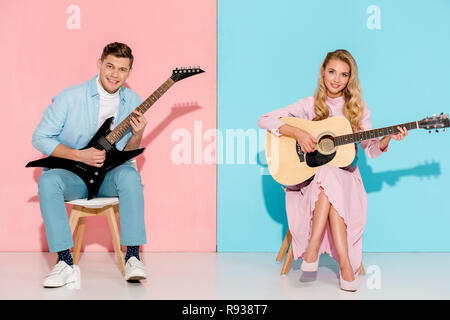 couple sitting on chairs, playing electric and acoustic guitars and looking at camera on pink and blue background