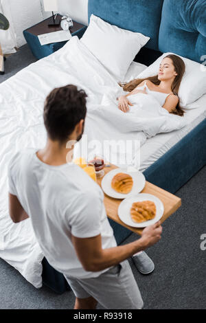 overhead view of man holding wooden tray with breakfast while woman sleeping in bed Stock Photo