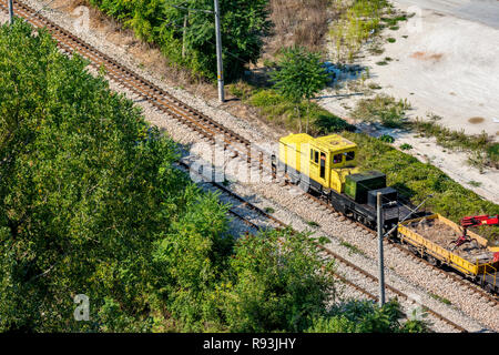 Yellow railway service diesel engine moving on rail line dragging wagons with pebbles and small red crane going to construction site. Rural Bulgarian  Stock Photo