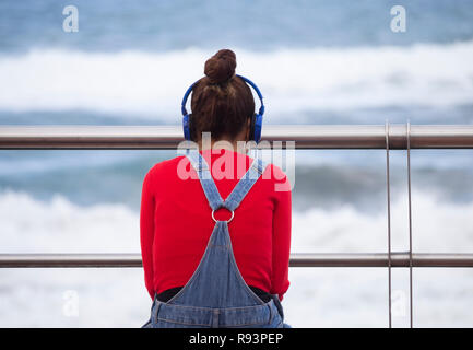 Rear view of young woman wearing headphones sitting on seat overlooking the sea. Stock Photo