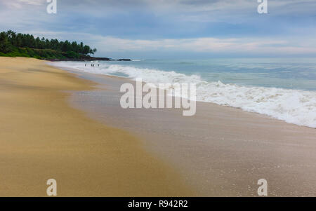 Coastline along sandy beach flanked by calm waters of Arabian Sea and a few palm tress and swimmers on horizon at dusk at Thottada, Kannur, Kerala. Stock Photo