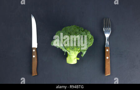 the concept of healthy eating. green, raw broccoli lying on a plate with instruments on a black background. healthy diet. green cabbage close-up. HLS. Stock Photo