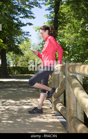 sporty adult woman with pink sweater, watching mobile smartphone leaning in wooden railing of footbridge, in park of Retiro, in Madrid, Spain. Vertica Stock Photo