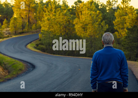 Active senior man stands alone on lonely road between mountains. Older man of back walking on lonely highway Stock Photo