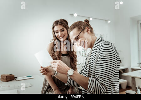 Long-haired designer in eyeglasses and a pretty model laughing at the picture Stock Photo