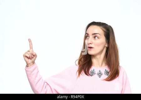 a beautiful and surprised young woman with dark hair looks away and she has an idea. in a pink jumper. Open mouth and pointing up. Isolated on white b Stock Photo