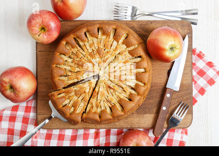 Homemade apple pie topped with slices of apples and cinnamon on wooden cutting board. Nearby are red apple, knife and checkered napkin. Top view. Stock Photo