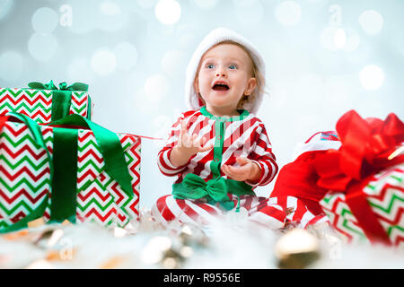 Cute baby girl 1 year old wearing santa hat posing over Christmas decorations with gifts. Sitting on floor with Christmas ball. Holiday season. Stock Photo