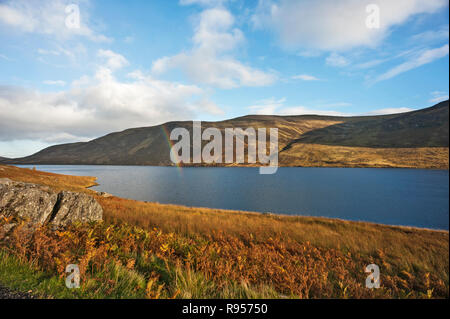 Partial rainbow over Loch Lee, Glen Esk, Angus, Scotland, UK, in foreground  on sunny day with loch side hills and bright cloudy blue sky in background. Stock Photo