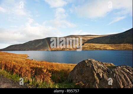 Partial rainbow over Loch Lee, Glen Esk, Angus, Scotland, UK, in foreground  on sunny day with loch side hills and bright cloudy blue sky in background. Stock Photo