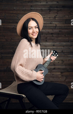 Attractive emotional young Caucasian woman in roll neck sweater and hat singing passionately and playing ukulele. Beautiful woman singer playing musical instrument indoors. People, music, joy and fun. Stock Photo