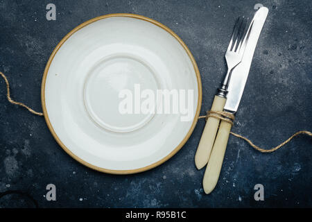 Empty white plate (ceramic, rustic) on a dark blue background with a knife and fork. Copy space. Stock Photo