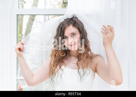 Closeup portrait of young female person, woman, bride in wedding dress, holding veil, hands, face, pearl necklace, standing by window, white curtains, Stock Photo