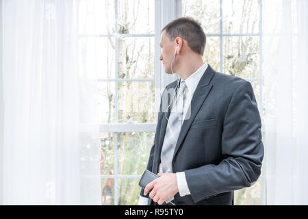 Closeup of young, happy smiling man, businessman, business suit, tie, standing by window in home, house room, white curtains, with earbuds headphones, Stock Photo