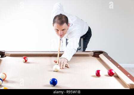 Young man, businessman in tie, white shirt, pants holding cue by pool table, playing snooker, billiard, billiards game, ready, striking white ball, ma Stock Photo