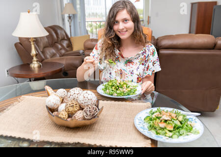 Young woman sitting with arugula, rocket, cucumber tempeh salad, holding fork, eating by glass table, tablecloths, decoration, decor in kitchen, livin Stock Photo