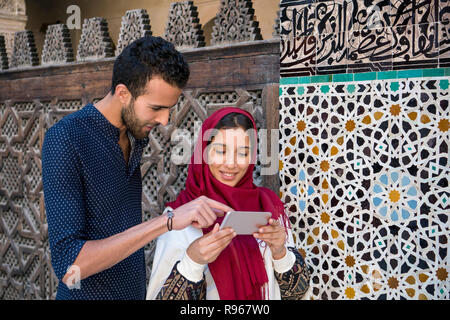 Smiling muslim couple looking at cellphone beside the arabesque decorated moroccan wall Stock Photo
