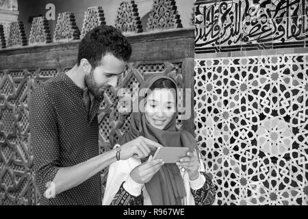 Black and white image of smiling muslim couple looking at cellphone beside the arabesque decorated moroccan wall Stock Photo
