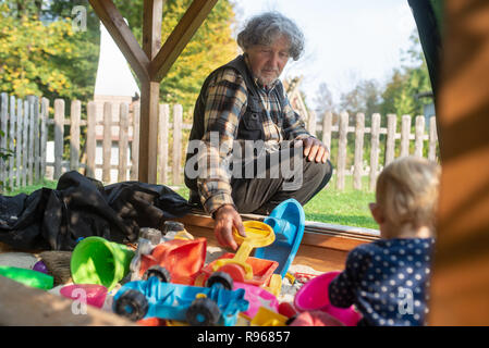 Grandfather playing with his granddaughter in a sand playground full of colorful toys on a sunny autumn day. Stock Photo